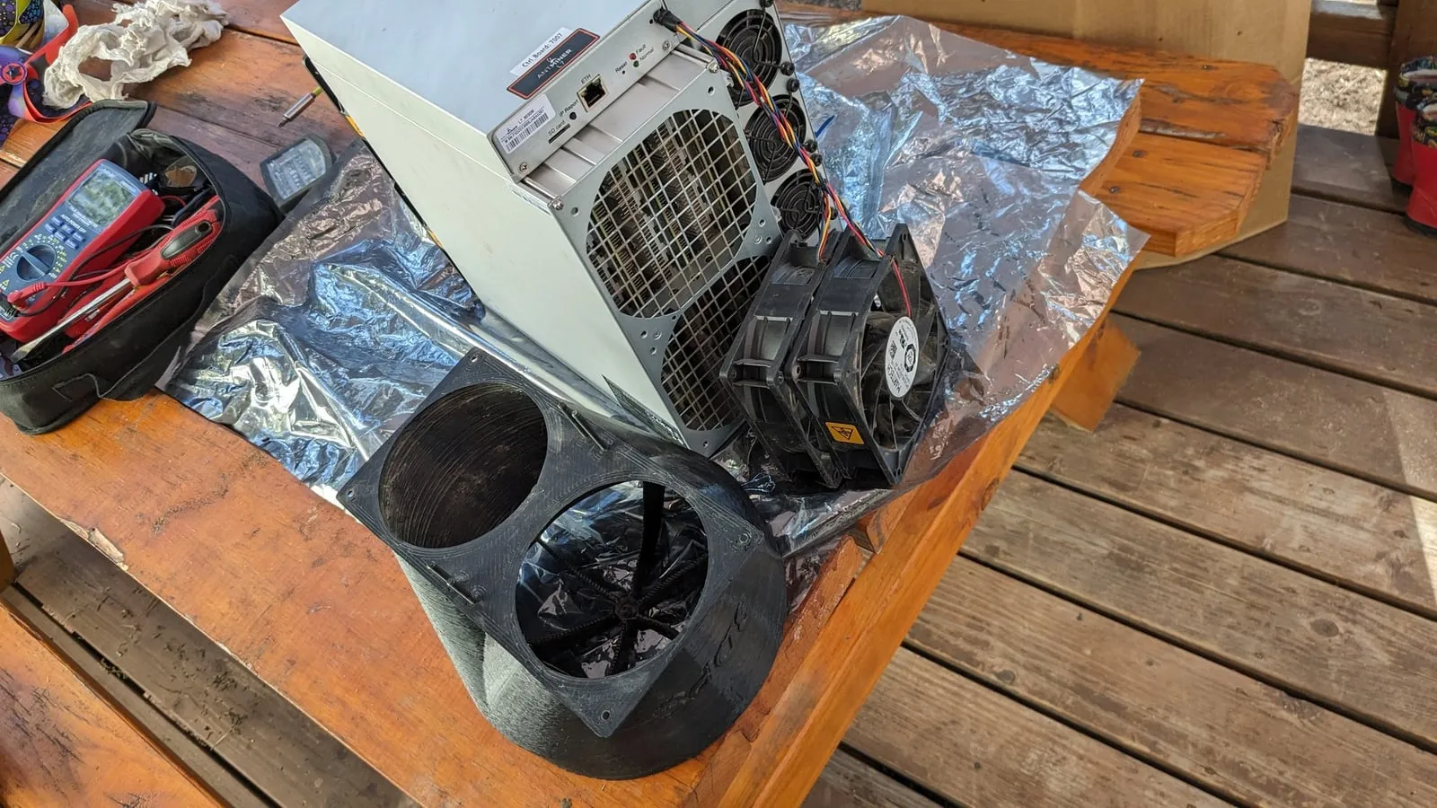Antminer L7 on a workbench next to the round black shroud that adapts its exhaust fans to the duct