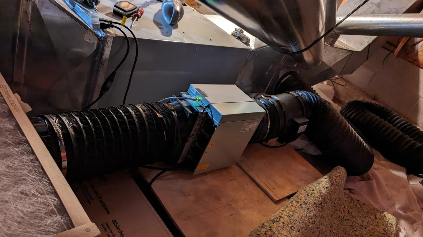The Antminer L7 in the crawl space, wired into a run of black flex duct between a filter-covered inlet on the left and the furnace plenum on the right