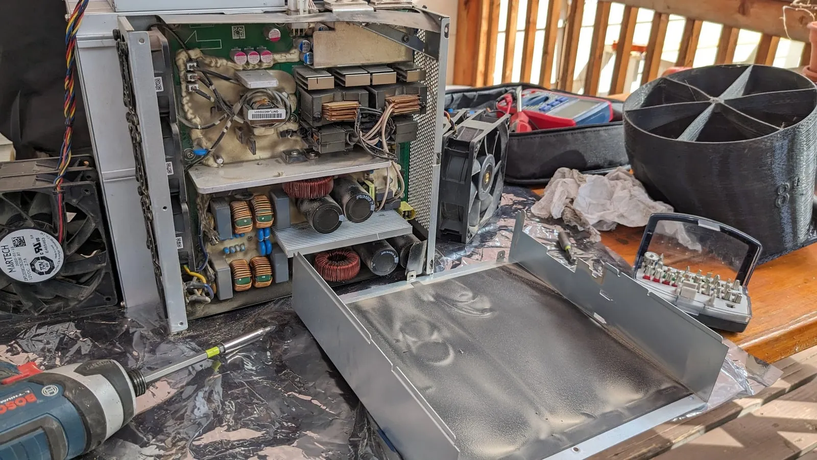 Antminer L7 opened up on an outdoor table, power supply and control board exposed, with the case lid and tools beside it